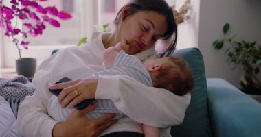 Anne bebeğini kanepede tutuyor, yüzü hem yorgunluk hem de sakinlik gösteriyor, anne olmanın günlük gerçekliğini yansıtıyor, rahat ve huzurlu bir yuvada.