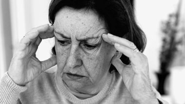 Şiddetli baş ağrısı ile mücadele eden yaşlı bir kadın, gözle görülür konsantrasyonda şakaklara masaj yapıyor. Yüz hatları ve yüz ifadeleri yoğun acıyı, zihinsel gerginliği, duygusal sıkıntıyı yansıtır.