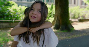 Kız kardeşi, güneşli bir günde erkek kardeşini sırtında taşıyor. Hem gülüyor, hem de bahçenin tadını çıkarıyor. Etrafı yeşillik ve gölgeli yürüyüş yolu ile çevrili.