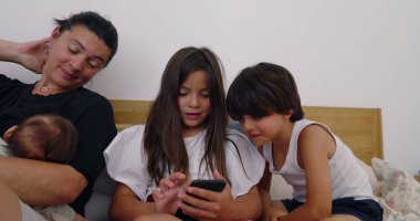 Anne kollarında bebek uyurken, kız kardeşiyle akıllı telefon kullanıyor, aile ile yatakta vakit geçiriyor, teknolojiyle meşgul çocuklar, rahat ev atmosferi, samimi bir bağ.