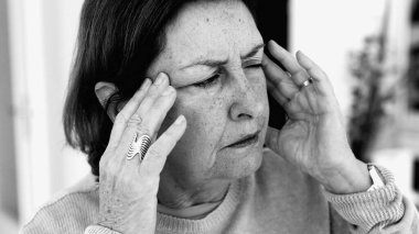 Şiddetli baş ağrısı ile mücadele eden yaşlı bir kadın, gözle görülür konsantrasyonda şakaklara masaj yapıyor. Yüz hatları ve yüz ifadeleri yoğun acıyı, zihinsel gerginliği, duygusal sıkıntıyı yansıtır.