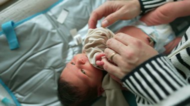Bebek bakıcı tarafından yumuşak bir paspasın üzerinde nazikçe giydiriliyor. Bebek sakince yukarı bakar. Bakıcının elleri elbiseyi özenle ve şefkatle ayarlar.