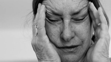 Yaşlı kadın yakın plan, gözleri kapalı, iki eli şakaklarına bastırıyor, derin kırışıklıklar ve acı dolu ifade, şiddetli stres ya da baş ağrısı