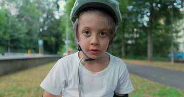 Parktaki koruyucu kıyafetlerle kaykayın üstünde oturan bir çocuk, başlığını takmış, konsantrasyonunu bozmadan ağaçlar ve yeşilliklerle çevrili, sakin, bulutlu bir günde.