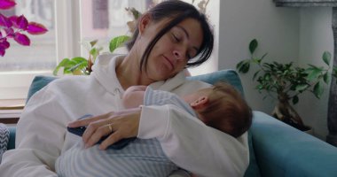 Anne bebeğini kucağında, bebek yüzüne dokunurken yavaşça kanepede dinleniyor. Huzurlu bir ev ortamında samimi bir yakınlık ve bağ anı.