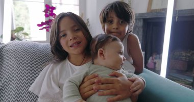 Kanepede bebekli kardeşler, kucağında bebek, pinpon topuyla komik surat yapan çocuk, aile bağları, rahat ev ortamı, neşeli anlar, kapalı mekan sahnesi