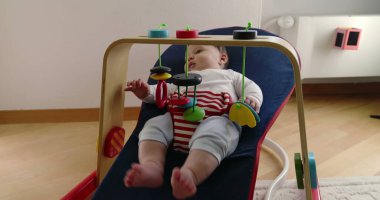 Bebek, bir fedainin içinde yatarken oyuncaklarla oynuyor, rahat bir ev ortamında merak ve neşe dolu bir an sergiliyor, çocukluk yıllarının masumiyetini ve neşesini vurguluyor.