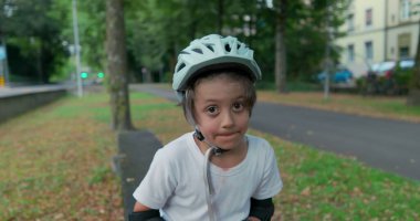 Kaykaylı bir kaykayın üzerinde oturan, düşünceli görünen, başlık takan, bulutlu bir günde ağaçlar ve yeşilliklerle çevrili bir çocuk.