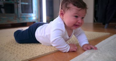 Ağlayan bebek yerde yatıyor, sıkıntı ve rahatsızlık gösteriyor, ev ortamında yumuşak bir paspas üzerinde duygularını ifade ediyor, erken gelişim zorluklarını ve hislerini vurguluyor.