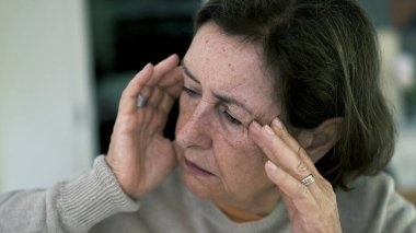 Başı kapalı, gözleri kapalı, şiddetli baş ağrısıyla boğuşan yaşlı bir kadın. Yüz ifadesi ve çatık kaşlar yoğun zihinsel ve duygusal baskı altında.