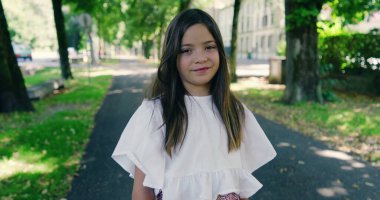 Parktaki ağaçlarla kaplı bir sokakta güler yüzlü bir kız, parlak bir günün tadını çıkarıyor, beyaz bir bluz ve desenli bir etek giyiyor, yemyeşil ile çevrili.