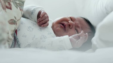 Yeni doğmuş bebek yatakta ağlıyor, rahatsız edici bir şekilde elini kaldırıyor. Bebeğin yüzünde sıkıntı var, ve jestler tedirginliği gösteriyor.