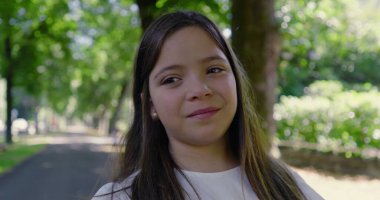 Parkta gülümseyen, beyaz bir bluz giyen, arka planda bulanık yeşillik ve ağaçlarla dolu uzun saçlı genç bir kızın yakın çekimi mutlu ve huzurlu bir anı yakalıyor.