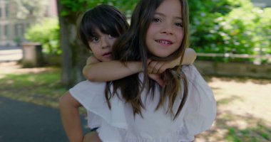 Kız kardeşi, güneşli bir günde erkek kardeşini sırtında taşıyor. Hem gülüyor, hem de bahçenin tadını çıkarıyor. Etrafı yeşillik ve gölgeli yürüyüş yolu ile çevrili.