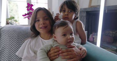 Kanepede bebekli kardeşler, kucağında bebek, pinpon topuyla komik surat yapan çocuk, aile bağları, rahat ev ortamı, neşeli anlar, kapalı mekan sahnesi