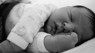 Yeni doğmuş bebeğe yakın plan. Yatakta huzur içinde uyuyor. Eli yüzünün yanında dinleniyor. Tek renkli bebeğin sakin ifadesi ve yumuşak, narin özellikleri.