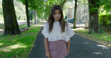 Mutlu kız, yeşil bir parkta, ağaçlarla kaplı bir yolda duruyor, kameraya sıcak bir şekilde gülümsüyor, beyaz bir bluz ve desenli bir etek giyiyor, güzel bir günün tadını çıkarıyor.