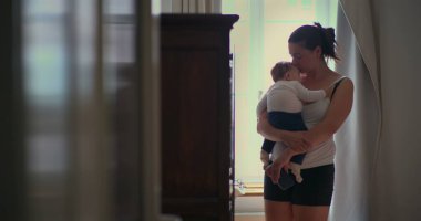 Anne bebeğini sevgi dolu bir şekilde kucağında tutuyor. Yumuşak ışıklı bir odada yürürken huzurlu bir bağ anı yaratıyor ve sıcak bir yuva kuruyor.