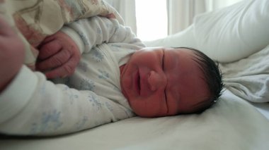 Yeni doğmuş bebek battaniyenin altında hareket ediyor, tuhaf bir ifadeyle yüzünü buruşturuyor, hayatın ilk ayındaki tipik hareketleri ve ifadeleri vurguluyor.