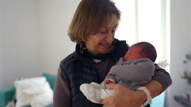 Büyükannem yeni doğmuş bir bebeği gururlu bir gülümsemeyle kucağında tutuyor, sıcak bir yerde duruyor, aile sevgisini ve çoklu-kuşak bağını gösteriyor.