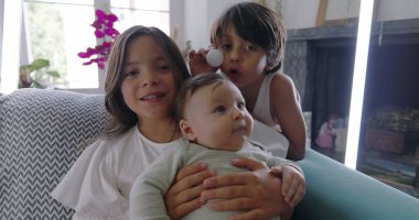 Kanepede bebekli kardeşler, kucağında bebek, pinpon topuyla komik surat yapan çocuk, aile bağları, rahat ev ortamı, neşeli anlar, kapalı mekan sahnesi