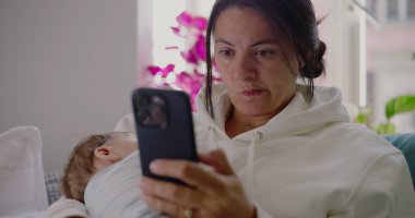 Anne bebeğini kucağında tutuyor ve rahat bir oturma odasında dinlenirken şüpheli bir şekilde telefonuna bakıyor. Camlardan içeri ışık süzülüyor. Sıradan bir aile anı.