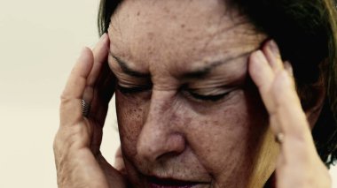 Yaşlı bir kadın şakaklarına sıkıca masaj yapıyor. Acıyı veya stresi azaltmaya odaklanıyor. Derin kırışıklıklar, yoğun yüz ifadesi, görülebilir gerilim şiddetli baş ağrısı iletir.