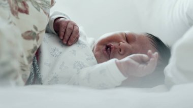 Yeni doğmuş bebek yatakta ağlıyor, rahatsız edici bir şekilde elini kaldırıyor. Bebeğin yüzünde sıkıntı var, ve jestler tedirginliği gösteriyor.