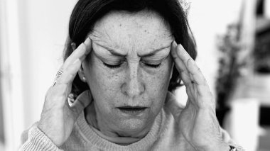 Şiddetli baş ağrısı ile mücadele eden yaşlı bir kadın, gözle görülür konsantrasyonda şakaklara masaj yapıyor. Yüz hatları ve yüz ifadeleri yoğun acıyı, zihinsel gerginliği, duygusal sıkıntıyı yansıtır.