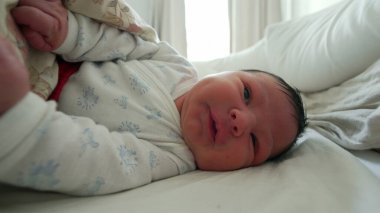 Yeni doğmuş bebek battaniyenin altında hareket ediyor, tuhaf bir ifadeyle yüzünü buruşturuyor, hayatın ilk ayındaki tipik hareketleri ve ifadeleri vurguluyor.