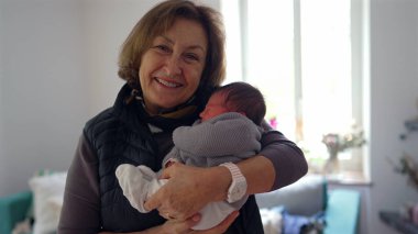 Büyükannem yeni doğmuş bir bebeği gururlu bir gülümsemeyle kucağında tutuyor, sıcak bir yerde duruyor, aile sevgisini ve çoklu-kuşak bağını gösteriyor.