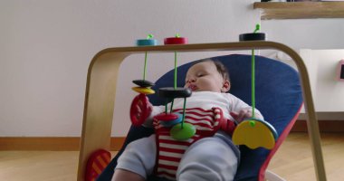 Bebek bir fedainin içinde uzanıyor ve asılı oyuncaklarla oynuyor, rahat bir ev ortamında keşiften ve oynamaktan zevk alıyor, çocukluk yıllarının merak ve masumiyetini vurguluyor.