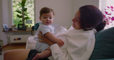 Anne, güneşli bir oturma odasında bebeğine gülümserken anne ve çocuk arasında sıcak ve sevgi dolu bir bağ kuruyor.