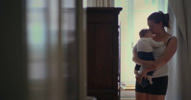 Anne bebeğini sevgi dolu bir şekilde kucağında tutuyor. Yumuşak ışıklı bir odada yürürken huzurlu bir bağ anı yaratıyor ve sıcak bir yuva kuruyor.