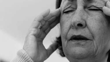 Yaşlı kadın yakın plan, gözleri kapalı, iki eli şakaklarına bastırıyor, derin kırışıklıklar ve acı dolu ifade, şiddetli stres ya da baş ağrısı