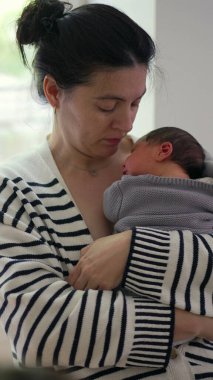 Anne yeni doğmuş bebeğini göğsüne yakın tutuyor, şefkatli bir ifadeyle aşağıya bakıyor. Anne ile çocuk arasındaki güçlü bağ sakin ve kapalı bir ortamda