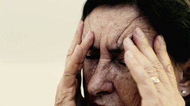 Yaşlı bir kadın şakaklarına sıkıca masaj yapıyor. Acıyı veya stresi azaltmaya odaklanıyor. Derin kırışıklıklar, yoğun yüz ifadesi, görülebilir gerilim şiddetli baş ağrısı iletir.