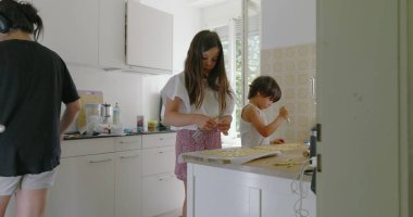 Kardeşler mutfakta birlikte yemek hazırlıyorlar, kız hamura odaklanıyor, oğlan asistanlığı, aile yemekleri, parlak mutfak, takım çalışması, anne arka planda, çekici bir an.