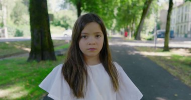 Parkta ağaçlarla kaplı bir yolda duran genç kız, kameraya tarafsız bir ifadeyle bakıyor, yeşil ve güneş ışığıyla çevrili, sakin bir açık hava anını yakalıyor.