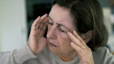 Başı kapalı, gözleri kapalı, şiddetli baş ağrısıyla boğuşan yaşlı bir kadın. Yüz ifadesi ve çatık kaşlar yoğun zihinsel ve duygusal baskı altında.