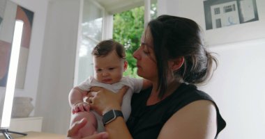 Anne sevgiyle bebeğini kucağında tutuyor, alınları birbirine değiyor, samimi ve duygusal bir bağ kurma anını paylaşıyor, sıcak bir yuva ortamında derin bir sevgi ve bağlantı kuruyor.