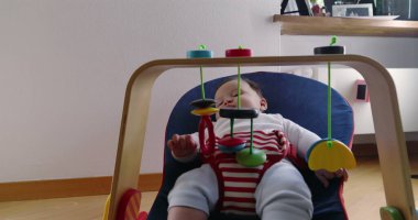 Bebek bir fedainin içinde uzanıyor ve asılı oyuncaklarla oynuyor, rahat bir ev ortamında keşiften ve oynamaktan zevk alıyor, çocukluk yıllarının merak ve masumiyetini vurguluyor.