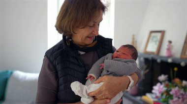 Büyükannem yeni doğmuş bir bebeği gururlu bir gülümsemeyle kucağında tutuyor, sıcak bir yerde duruyor, aile sevgisini ve çoklu-kuşak bağını gösteriyor.