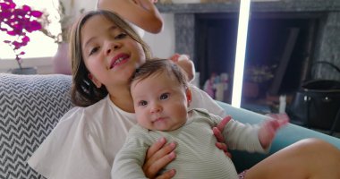 Bebek kardeşi koltukta otururken kucağında, bebek merakla yukarıyı seyrediyor, sıcak bir ev ortamında şefkatli ve huzurlu bir aile anı yaratıyor.