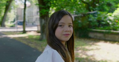 Parkta ağaçlarla kaplı bir yolda duran genç kız, kameraya tarafsız bir ifadeyle bakıyor, yeşil ve güneş ışığıyla çevrili, sakin bir açık hava anını yakalıyor.