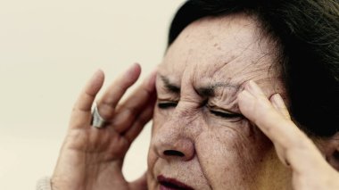 Yaşlı bir kadın şakaklarına sıkıca masaj yapıyor. Acıyı veya stresi azaltmaya odaklanıyor. Derin kırışıklıklar, yoğun yüz ifadesi, görülebilir gerilim şiddetli baş ağrısı iletir.