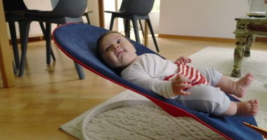 Bebek bir fedainin içinde uzanıyor, gülümsüyor ve içerideymiş gibi görünüyor, rahat bir ev ortamında bir anlık rahatlığın tadını çıkarıyor, erken çocukluğun neşesini ve masumiyetini vurguluyor.