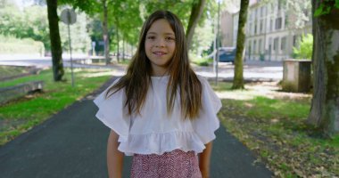 Uzun saçlı bir kız ileri doğru yürüyor ve sonra geri dönüp kameraya bakarken ağaçlarla kaplı patikaları ve arka planda binaları olan güneşli bir parkta yürüyor.