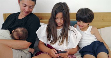 Anne sütü emziren bebek, kız kardeşinin yanında akıllı telefon tutması, aile paylaşımı anı yatakta, teknoloji ve bağ kurma, rahat bir yuva kurma, sıradan bir gün.