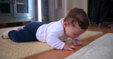 Ağlayan bebek yerde yatıyor, sıkıntı ve rahatsızlık gösteriyor, ev ortamında yumuşak bir paspas üzerinde duygularını ifade ediyor, erken gelişim zorluklarını ve hislerini vurguluyor.